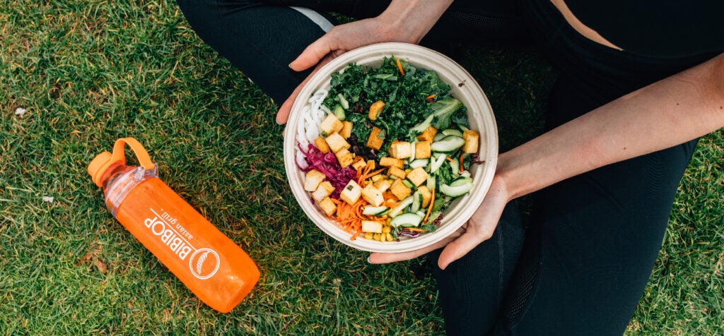 A person sitting on the grass holding a BIBIBOP bowl in their lap with a BIBIBOP Asian Grill water bottle next to them