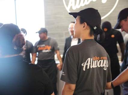 The back of a BIBIBOP Asian Grill employee standing in a team huddle wearing a BIBIBOP shirt saying: Take care always