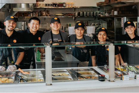 Six BIBIBOP Asian Grill employees standing behind a Build Your Own BIBIBOP Bowl restaurant counter smiling at the camera