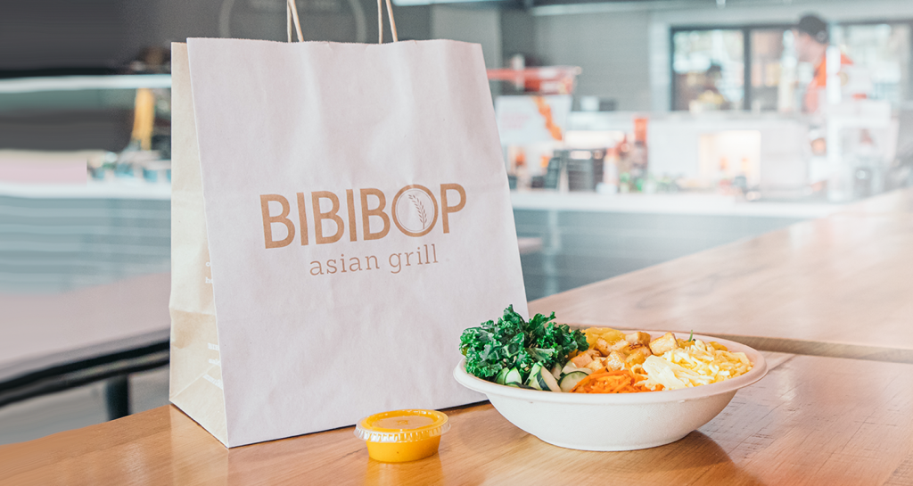 A BIBIBOP Asian Grill takeout bag sitting on a counter next to a BIBIBOP bowl and container of sauce