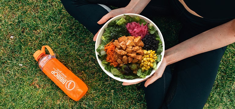 A person sitting on the grass holding a BIBIBOP bowl in their lap with a BIBIBOP Asian Grill water bottle next to them