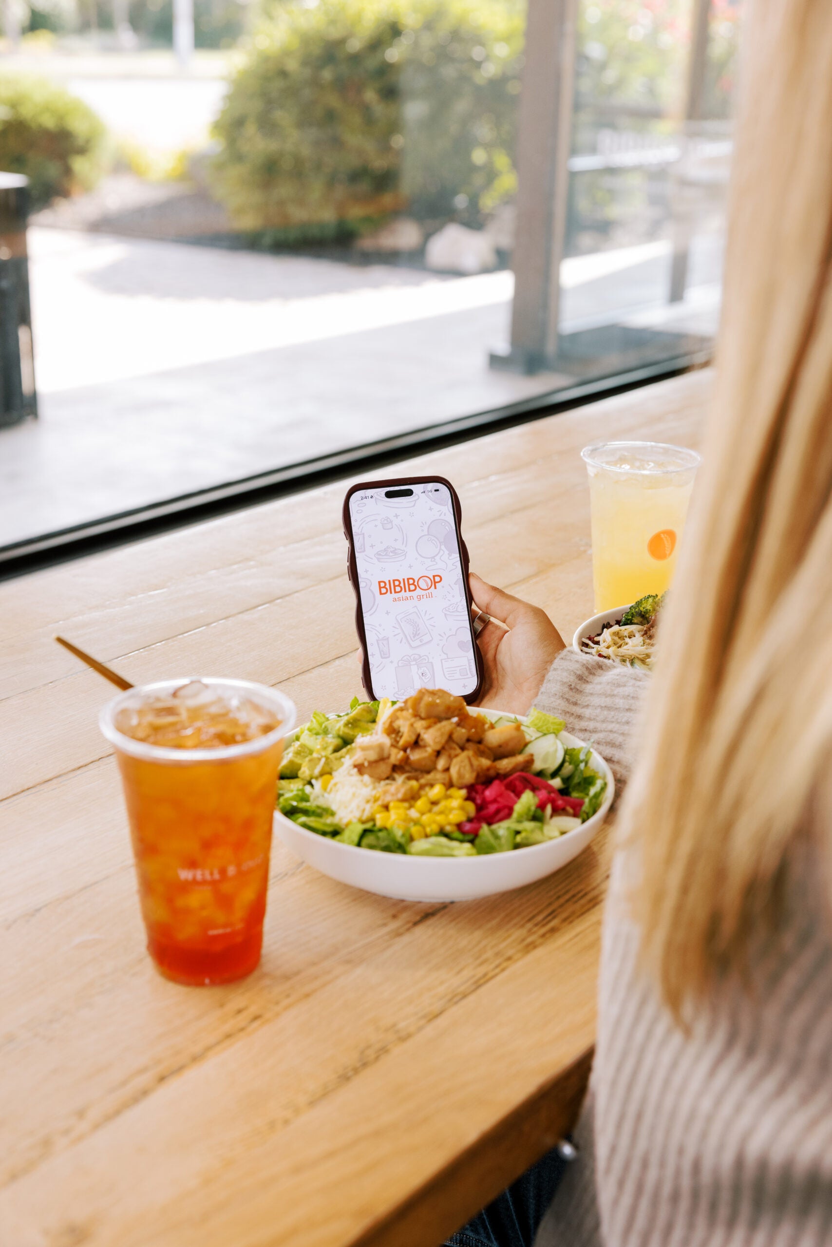 Woman holding phone with Bibibop app and bowl of rice with protein with drink