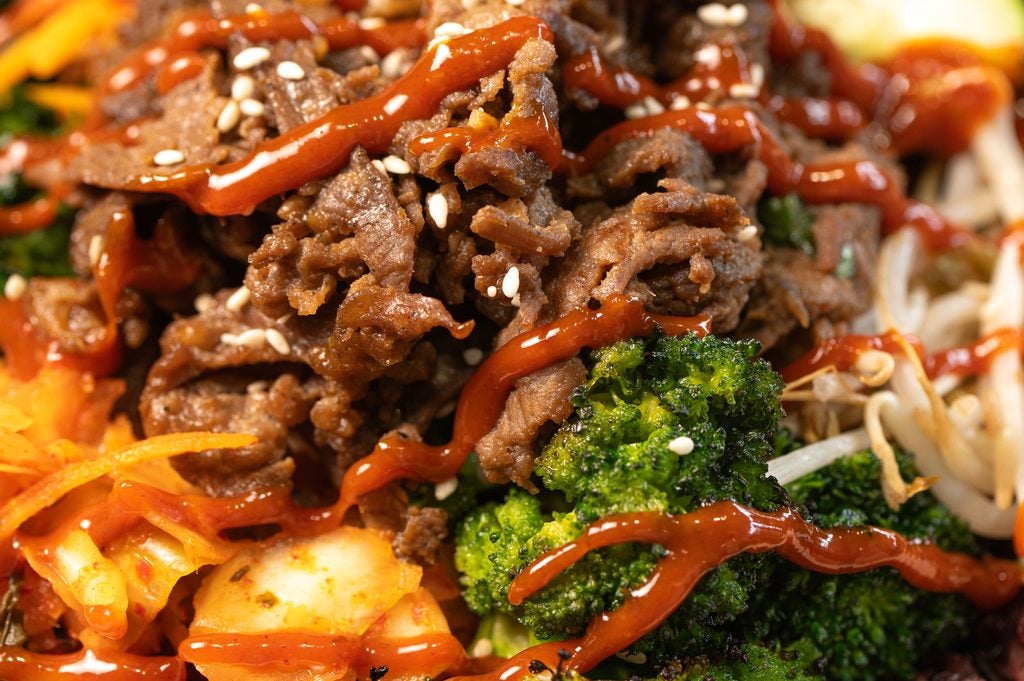 Close up of a BIBIBOP bowl with Korean BBQ Beef, Roasted Sesame Broccoli, Bean Sprouts and Kimchi under a drizzle of sauce